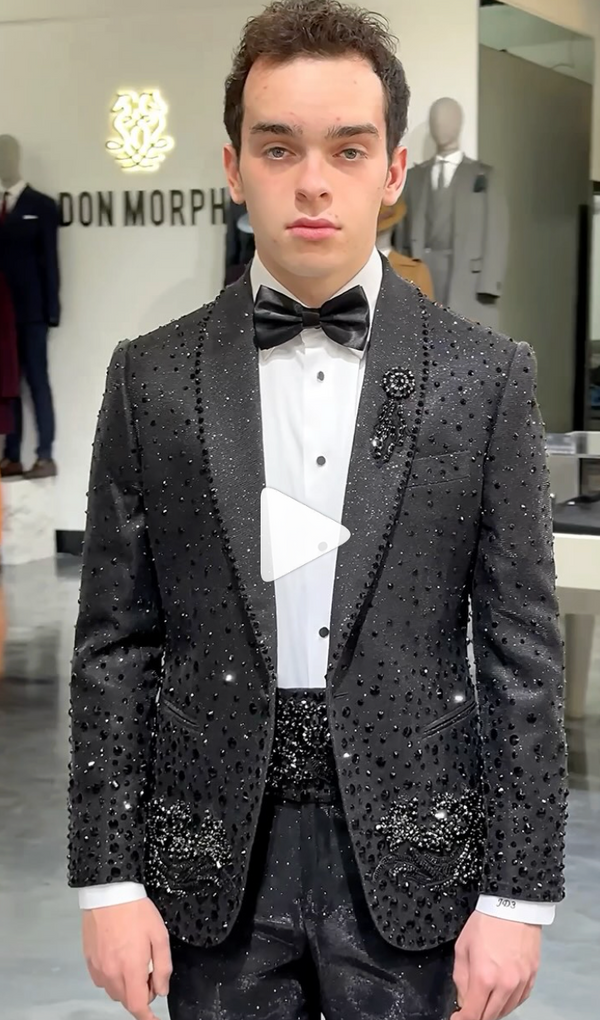 Stylish young man in detailed black tux stands inside shop with firm expression